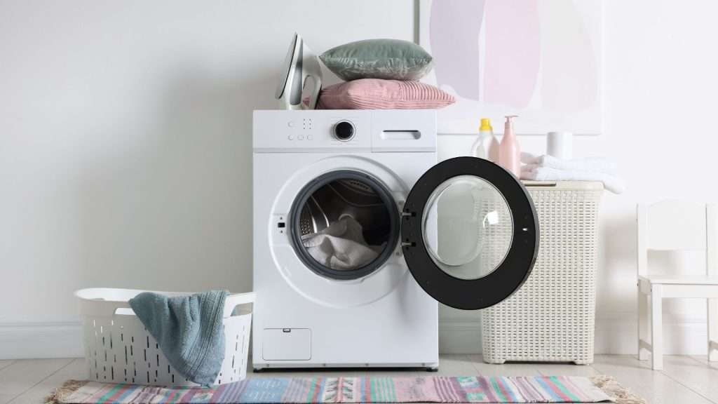 Front-loading washing machine with door open in a bright laundry room, surrounded by a laundry basket, folded towels, detergent bottles, and a woven hamper.